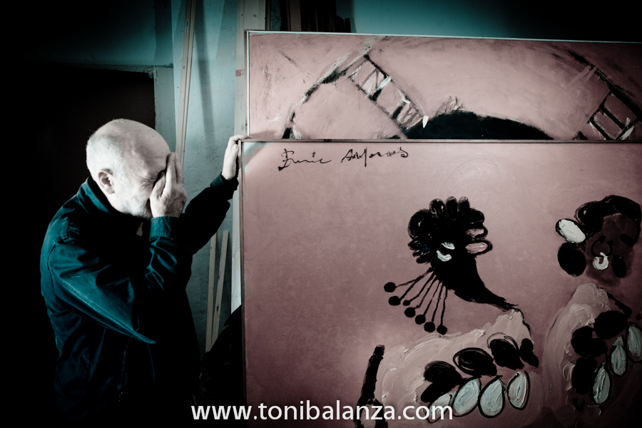 El pintor Enric Alfons, se tapa la cara a modeo de máscara con su propia mano, junto a una de sus obras en Valencia. Fotografía de Toni Balanzà